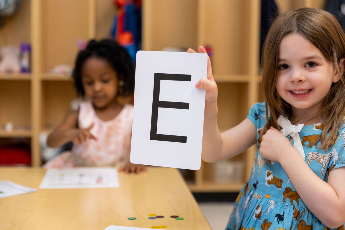 Young student at the New E3 School holding up a flash card with the letter E on it.